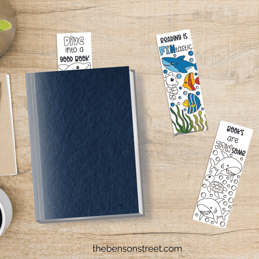 A book with a shark bookmark holding the page on a desktop with two other shark bookmarks on the table.