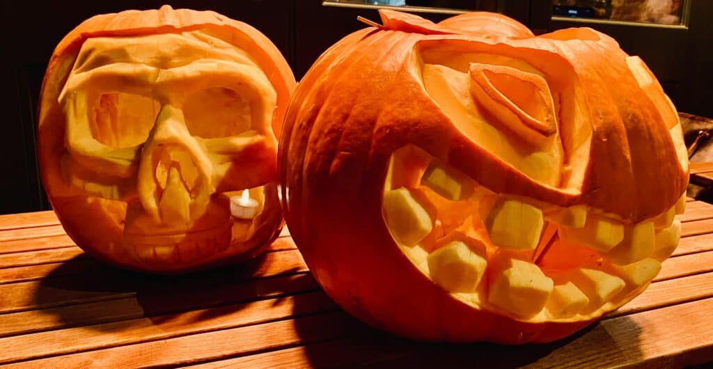 Two detailed pumpkin carvings with three dimensional elements, one a classic jack-o'lantern with teeth and eyes and the other a skull.