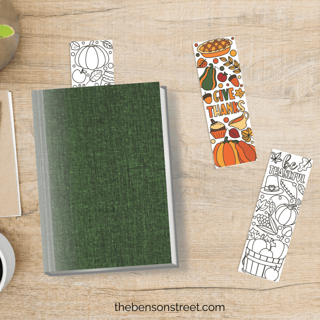 A desk shot from overhead with a book and a bookmark in it, and on the table next to it is a bookmark that says give thanks colored in, and another that says be thanksful ready to be colored in.
