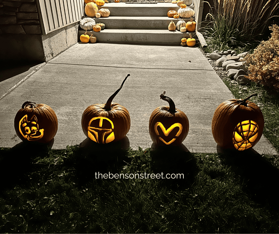 Four carved pumpkins lit up at night with Bluey, the Mandalorian, a Heart, and the Amazing Spiderman all in a row.